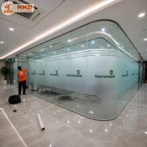 Thi công decal mờ văn phòng in logo Vietcombank trên vách kính cong. Decal mờ kết hợp logo nổi bật giúp phân chia không gian hiệu quả, tạo sự riêng tư và mang lại vẻ đẹp sang trọng cho văn phòng.