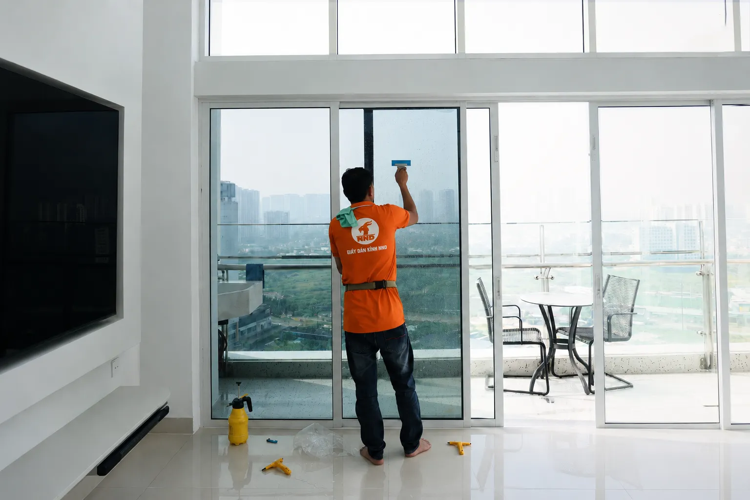 Kỹ thuật viên NND dùng gạt hoàn thiện phim cách nhiệt trên cửa kính, bên dưới có bình xịt và dụng cụ thi công.