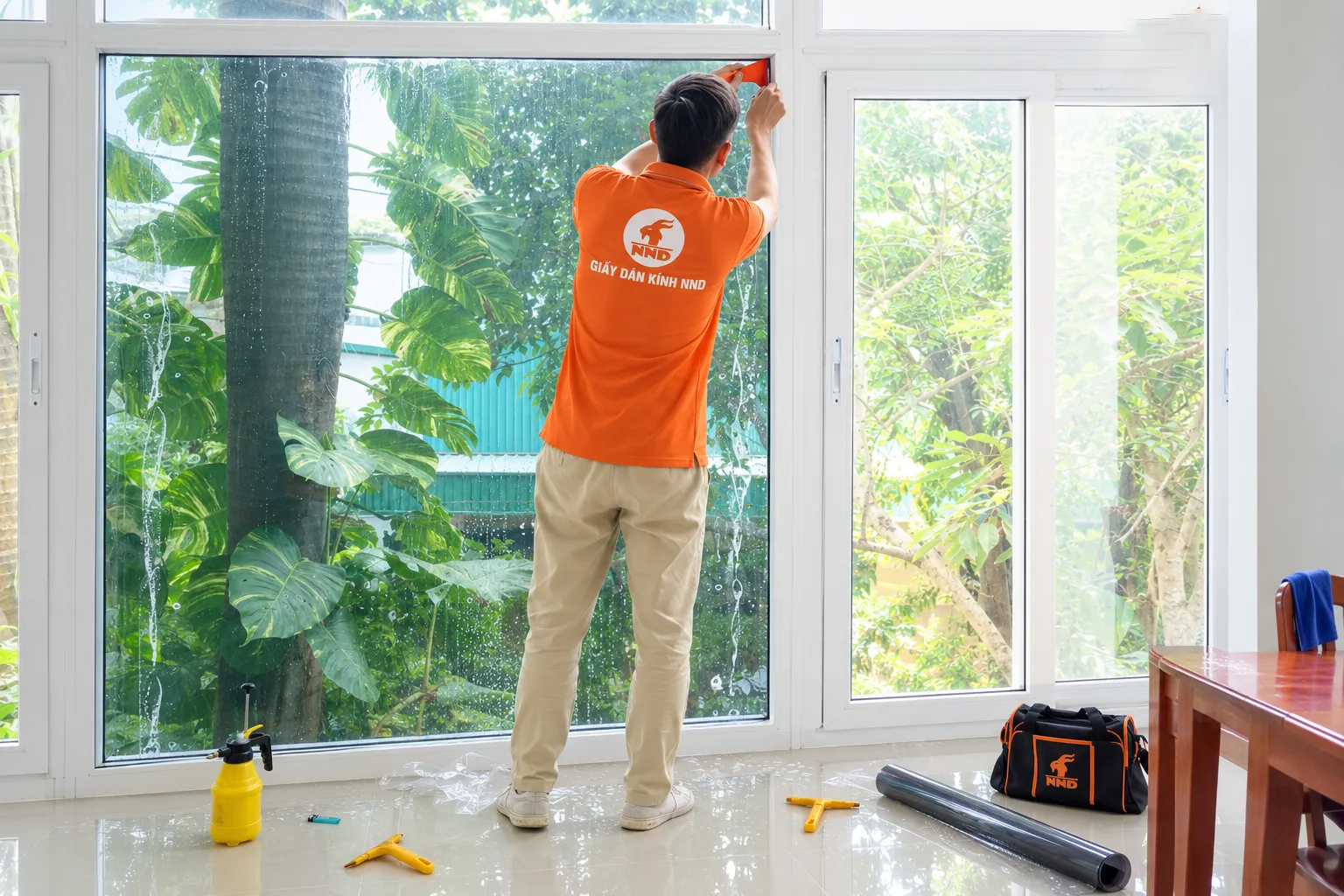 Thợ NND xử lý mép phim cách nhiệt trên ô cửa kính lớn, cạnh đó có cuộn phim và bộ dụng cụ thi công.