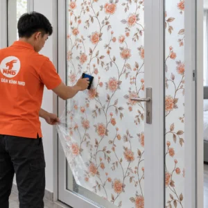 Thợ NND áo cam giữ mép tấm và dùng gạt dán A55 hoa dây trên cửa kính cạnh phòng ngủ